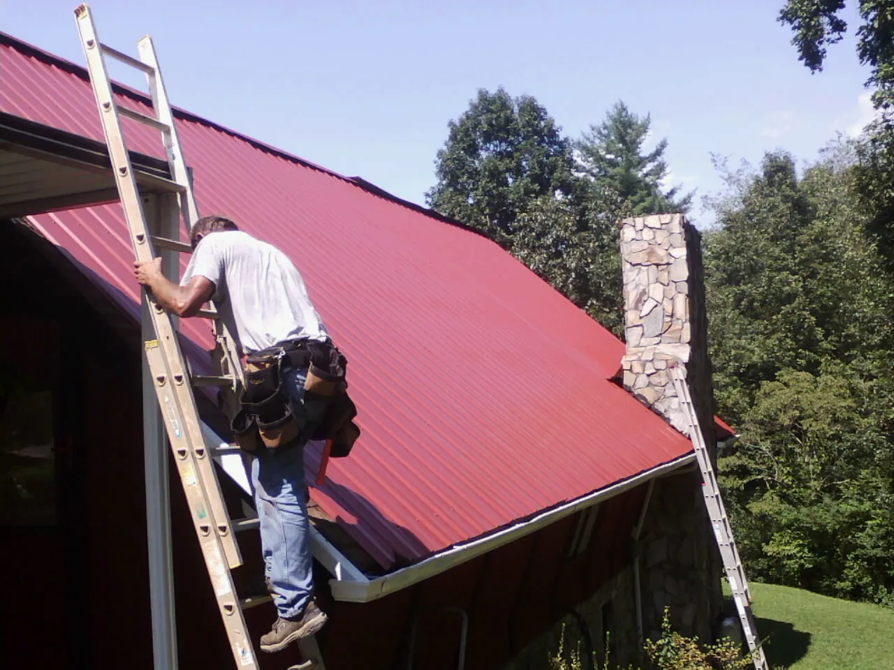 Roofer on a ladder beside a red metal roof and stone chimney — Soffit & Fascia Repair by Roofing Brothers in Elkins Park